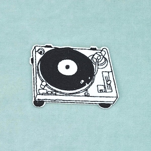 May include: A black and white embroidered patch featuring a detailed line art illustration of a turntable. The design shows a circular record on a rectangular base with a tonearm, control knobs, and sliders, rendered in high-contrast black thread.