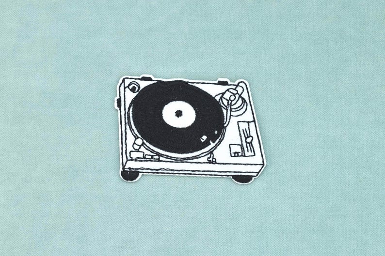 May include: A black and white embroidered patch featuring a detailed line art illustration of a turntable. The design shows a circular record on a rectangular base with a tonearm, control knobs, and sliders, rendered in high-contrast black thread.