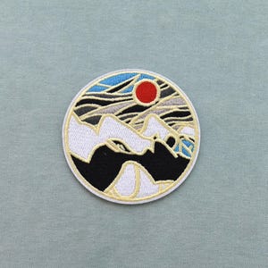 Peut inclure: Un écusson brodé circulaire présentant un paysage avec un soleil rouge vif, des sommets enneigés blancs, des bases de montagnes noires et des accents de ciel bleu, le tout entouré d'une fine bordure circulaire de couleur claire.