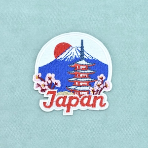 Peut inclure: Un écusson brodé circulaire représentant une montagne bleue au sommet enneigé blanc, un soleil rouge, une pagode rouge à plusieurs niveaux et des fleurs de cerisier roses. Le mot Japan est brodé en lettres cursives rouges en bas.