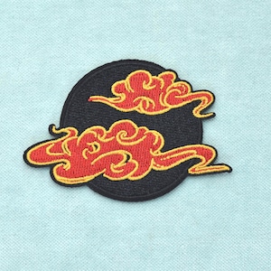 May include: A circular embroidered patch featuring a solid black background with two stylised red clouds outlined in vibrant yellow thread. The design showcases intricate stitching details that define the swirling shapes of the clouds.