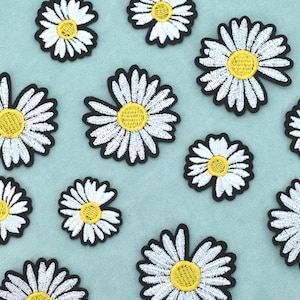 May include: A collection of embroidered daisy patches featuring white petals and yellow centres, each outlined with a thick black border. The patches vary in size and are arranged in a scattered pattern, showcasing detailed stitching throughout.
