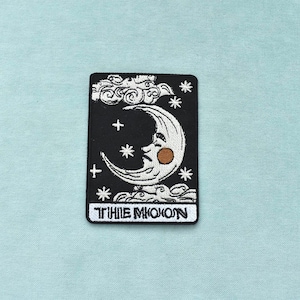 Pode incluir: Patch bordado com uma lua crescente com um rosto, estrelas e nuvens sobre um fundo preto. O patch tem as palavras "THE MOON" em branco na parte inferior. O design é em branco, castanho e preto.