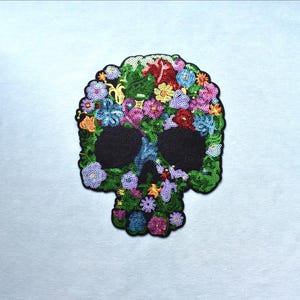 Peut inclure: Un écusson en forme de crâne recouvert de sequins multicolores vibrants. Le motif présente une disposition dense de fleurs dans des tons de vert, rouge, bleu, jaune et violet, avec deux grandes orbites circulaires en noir uni.
