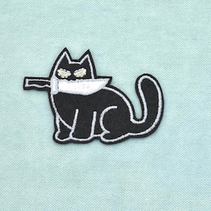 May include: An embroidered patch featuring a black cat with white outlines, sitting in profile with a long tail. The cat holds a silver-colored knife with a black handle in its mouth, and its eyes are depicted with small, light-colored circular details.