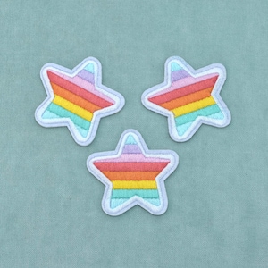May include: Three embroidered star-shaped patches featuring horizontal stripes in a rainbow colour palette of light purple, pink, red, orange, yellow, green, and light blue. Each star has a thick, light-coloured stitched border around the edges.