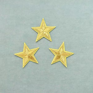 Puede incluir: Tres parches idénticos en forma de estrella de cinco puntas con bordado de hilo dorado metálico, un patrón geométrico cosido distintivo en el centro y un borde dorado sólido, dispuestos en formación triangular sobre una superficie plana.