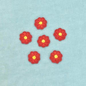 Può includere: Cinque piccole applicazioni floreali all'uncinetto di forma circolare disposte a pentagono. Ogni fiore presenta petali rossi vivaci con bordi smerlati che circondano un centro testurizzato di colore giallo brillante.