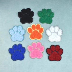 Op de afbeelding: Acht geborduurde pootafdruk-patches gerangschikt in een raster. De kleuren zijn zwart, groen, lichtblauw, oranje, wit, donkerblauw, roze en donkerrood. Elke patch heeft een getextureerd oppervlak met een glinsterende rand rond de pootvorm.