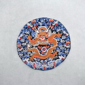 Könnte beinhalten: Runder, gestickter Aufnäher mit einem orangefarbenen Drachen auf schwarzem Hintergrund. Der Drache ist von blauen, weißen und roten Wolkenmustern umgeben. Der Aufnäher ist wahrscheinlich für dekorative Zwecke bestimmt.