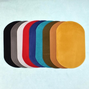 Op de afbeelding: Een collectie van negen ovaalvormige stoffen matten gerangschikt in een rij, met een spectrum aan kleuren waaronder zwart, grijs, wit, rood, blauw, taling, olijfgroen, bruin en mosterdgeel, elk met een glad, uniform en effen gekleurd oppervlak.