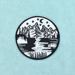 Può includere: Una toppa ricamata circolare con un design paesaggistico in bianco e nero. La scena raffigura montagne frastagliate, una luna crescente e stelle sopra un lago riflettente circondato da pini e fitto fogliame boschivo con bordo nero.