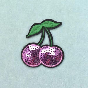 Puede incluir: Un parche termoadhesivo con forma de cerezas compuesto por dos frutos redondos cubiertos de lentejuelas rosas brillantes. El parche tiene un borde bordado en negro, dos hojas bordadas en verde y tallos verdes que unen las hojas a los frutos.