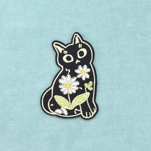 Peut inclure: Un écusson brodé représentant un chat noir aux grands yeux blancs et à la petite bouche. Le chat est orné de deux fleurs de marguerite blanches à cœur jaune et de feuilles vertes sur le corps, le tout bordé d'un fin liseré blanc sur fond noir.