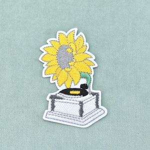 May include: An embroidered patch featuring a white gramophone with a black record on the turntable. A large, bright yellow sunflower with a grey center serves as the gramophone horn, connected by a curved green stem to the base of the device.