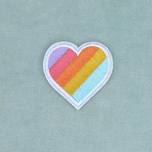 May include: A heart-shaped embroidered patch featuring diagonal stripes in lavender, pink, orange, yellow, and light blue. The patch has a white stitched border and a textured fabric finish, creating a clean and defined shape for application.