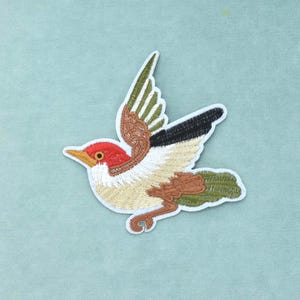 Pode incluir: Um emblema bordado apresentando um pássaro em voo com cabeça vermelha brilhante, bico amarelo e peito branco. As asas exibem uma mistura de pontos castanhos, verde-azeitona e pretos, enquanto as penas da cauda são de um verde-azeitona texturizado.
