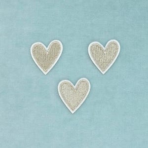 Può includere: Tre toppe a forma di cuore con una finitura in filo metallico dorato testurizzato. Ogni cuore è delineato da un bordo bianco netto, creando un contrasto distinto con il centro dorato scintillante dei pezzi in tessuto ricamato.