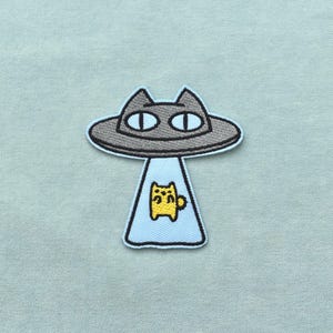 Peut inclure: Un écusson brodé représentant un OVNI en forme de chat gris avec de grands yeux blancs et un contour noir. Un rayon tracteur bleu clair descend du vaisseau, contenant une petite créature jaune à quatre pattes avec un contour noir simple.