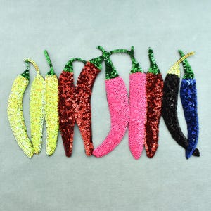 Peut inclure: Une collection d'écussons de piments recouverts de sequins de différentes couleurs : jaune, rouge, rose, noir et bleu. Chaque écusson a une tige verte et est disposé sur un fond gris clair.