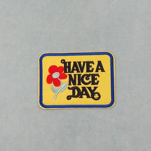 Peut inclure: Patch brodé jaune avec une bordure bleue. Le patch représente une fleur rouge avec une tige verte et le texte "HAVE A NICE DAY" en noir.