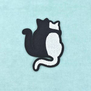 May include: An embroidered patch featuring two cats sitting back-to-back. One cat is solid black and the other is solid white. The design shows the cats in profile with their tails curved, creating a cohesive silhouette against a light blue background.