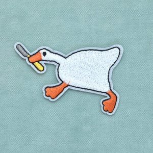 Op de afbeelding: Een geborduurde strijkpatch met een witte gans met oranje voeten en een oranje snavel. De gans is afgebeeld in een rennende houding en houdt een klein zilverkleurig mes met een geel handvat in de snavel tegen een lichtblauwe achtergrond.