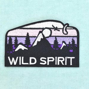 Op de afbeelding: Een rechthoekige geborduurde patch met afgeronde bovenhoeken met een berglandschap bij nacht. Het ontwerp bevat een zwart bergsilhouet, dennenbomen, een maan en een vogel tegen een paarse en witte lucht met de tekst WILD SPIRIT.