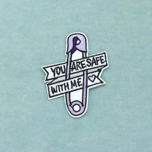 Pode incluir: Um emblema bordado com um alfinete de segurança roxo claro orientado verticalmente. Uma faixa branca com bordas pretas e o texto preto YOU ARE SAFE WITH ME está sobreposta ao alfinete, acompanhada por um pequeno ícone de coração roxo à direita.
