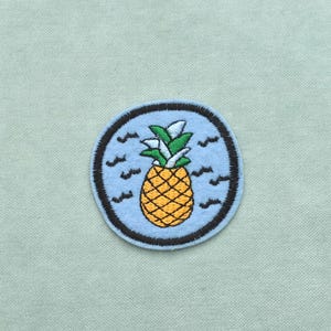 Peut inclure: Écusson brodé représentant un ananas jaune avec des feuilles vertes sur fond bleu. L'ananas est entouré d'une bordure noire circulaire, avec des vagues stylisées. L'écusson est sur un tissu vert clair. Environ 6 cm de diamètre.