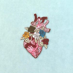 Peut inclure: Un écusson brodé représentant un cœur anatomique dans des tons de rose et de rouge, orné d'un bouquet de petites fleurs jaunes, blanches, bleues et rouges avec des feuilles vertes, le tout bordé d'un contour blanc sur fond bleu clair.