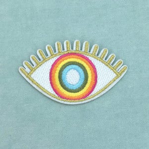 Op de afbeelding: Een geborduurde patch in de vorm van een oog met een regenboogkleurige iris bestaande uit concentrische cirkels in lichtblauw, groen, geel, oranje en roze. Het oog heeft goudkleurige geborduurde wimpers en een witte achtergrond met een gouden rand.