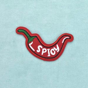 Op de afbeelding: Een geborduurde patch in de vorm van een rode chilipeper met een groene steel. Het woord SPICY is in witte hoofdletters in het midden van de peper geborduurd. De patch heeft een rode rand en ligt op een lichtblauwe stoffen achtergrond.