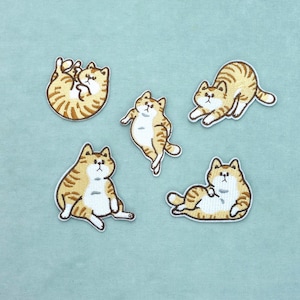 Pode incluir: Cinco emblemas bordados apresentando gatos malhados laranja em várias poses. Cada gato tem pelo branco no peito e na barriga, com listras castanhas. Os gatos são mostrados deitados, sentados, a espreguiçar-se e a rebolar.