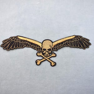 May include: Embroidered patch depicting a skull with gold wings and crossed bones. The wings are black with gold detailing. The patch is designed for application to clothing or accessories, and is likely made of fabric.