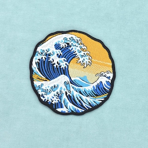 Puede incluir: Un parche bordado circular que presenta un diseño de ola oceánica estilizada con detalles en hilo azul oscuro y blanco sobre un fondo amarillo dorado, acabado con un borde grueso cosido en negro alrededor del contorno.