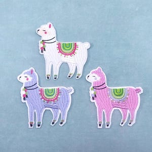 May include: Three embroidered llama patches in white, lavender, and pink. Each llama has a colourful saddle blanket and a decorative neck piece. The patches are on a light blue background.