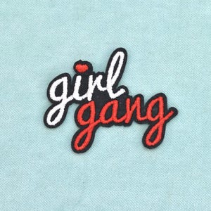 May include: An embroidered patch featuring the text girl gang in a cursive script. The word girl is stitched in white thread with a small red heart above the letter i, while the word gang is stitched in red thread. The entire design has a black border.