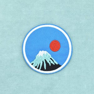 以下が含まれることがあります： 青い空の背景、鮮やかな赤い太陽、白い雪を頂いた山頂と黒い山裾を持つ山が描かれた円形の刺繍ワッペン。全体が太い白い縁取りで囲まれており、水色の質感のある布地の上に配置されています。