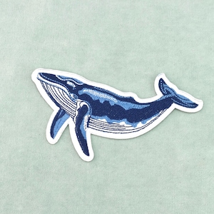 Peut inclure: Écusson brodé bleu et blanc représentant une baleine. La baleine est représentée dans des tons de bleu avec des coutures détaillées, sur un fond bleu clair. L'écusson a une bordure blanche.