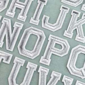 Peut inclure: Une collection de patchs alphabet thermocollants présentant des lettres argentées métalliques avec une bordure blanche. Chaque lettre est conçue dans un style de police bloc universitaire classique, disposée en rangées sur un tissu vert clair.