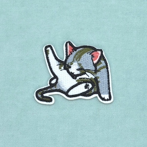 May include: An embroidered patch featuring a grey and white tabby kitten in a seated position, grooming its raised hind leg. The kitten has pink inner ears, dark grey stripes, and a white belly, all outlined in black thread on a white border.