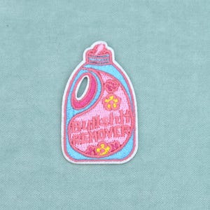 Peut inclure: Un écusson brodé en forme de bouteille de lessive avec une bordure bleu clair et un centre rose. Le motif présente des accents de fleurs jaunes et le texte BULLSHIT REMOVER brodé en fil rose sur la surface avant de l'objet.