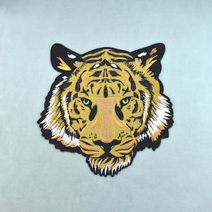 Può includere: Una toppa ricamata a forma di testa di tigre con dettagli in fili giallo oro, nero e bianco. La tigre ha occhi verdi penetranti, un naso testurizzato e strisce nere sul viso, il tutto delineato da un bordo nero spesso.