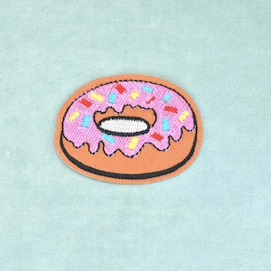 May include: An embroidered patch shaped like a doughnut with a tan base, pink icing, and small rectangular sprinkles in yellow, blue, and red. The doughnut has a dark brown outline and a white centre hole, featuring dense stitching throughout the design.