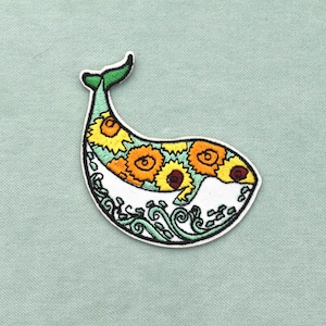 Peut inclure: Un écusson brodé en forme de baleine, présentant un corps recouvert de motifs de tournesols jaunes et orange, une queue verte et un ventre blanc avec des motifs de vagues vertes stylisées, le tout bordé d'un fil noir sur un contour blanc.