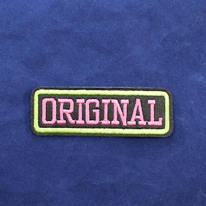 Peut inclure: Un écusson rectangulaire avec le mot "ORIGINAL" brodé en lettres roses. Le texte est souligné en noir et encadré par une bordure vert néon. L'écusson est posé sur un fond bleu foncé.