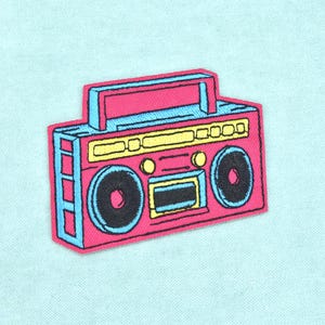 Könnte beinhalten: Ein gestickter Aufnäher in Form eines Retro-Boombox-Radios. Das Design zeigt einen leuchtend pinken Körper mit hellblauen Akzenten, gelben Bedienfeldern und zwei großen schwarzen, runden Lautsprechern. Das Motiv hat einen pinken Rand.