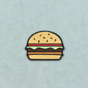May include: An embroidered patch shaped like a hamburger with a tan bun, a red tomato slice, light green lettuce, a brown meat patty, and a yellow cheese slice, all outlined in black thread against a light blue textured fabric background.