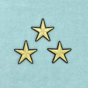 May include: Three embroidered star patches arranged in a triangular formation. Each star features a metallic gold thread finish with a distinct black stitched border, creating a sharp contrast against the light blue fabric background.